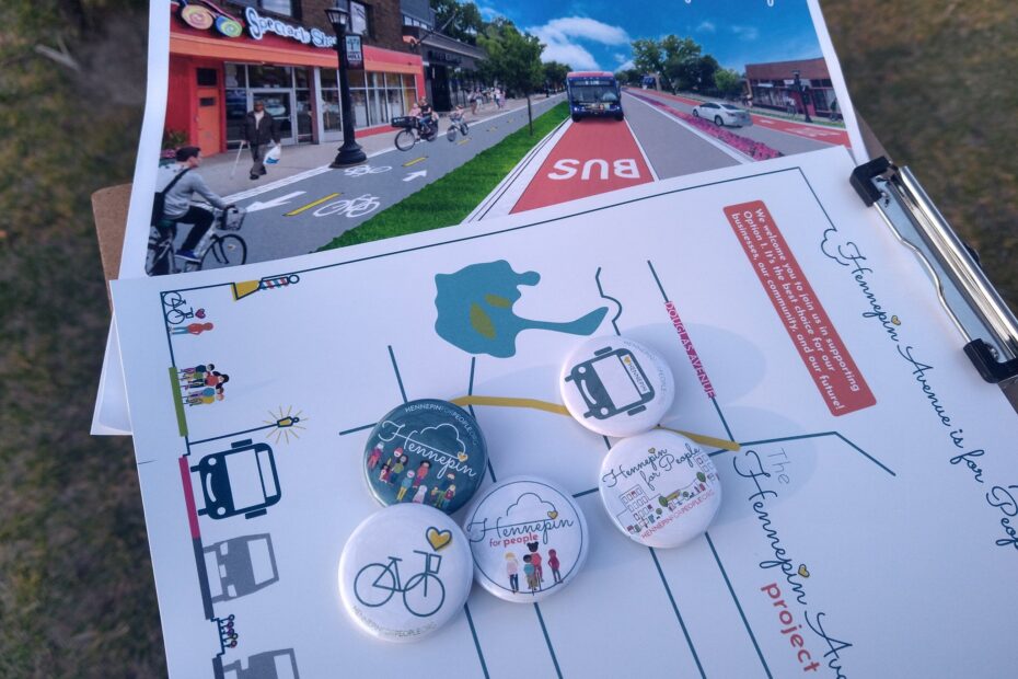 Clipboard with Hennepin for People outreach materials. Image of future street, map of project area, and buttons. Smiley bus face, Streetscape, Kids on wheels (biking, skateboarding, scooter), bike with yellow heart above, and our logo our people involved with Hennepin for People.