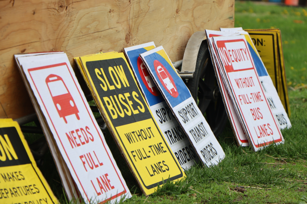 Several signs: Bus Needs Full Lane, Slow Buses Without Full-Time Lanes, Support Transit Riders, No Equity Without Full-Time Bus Lanes, Don't Delay The Bus.