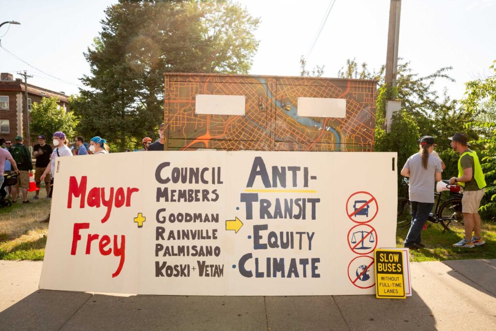 Hennepin for People sign: Mayor Frey + Council Members Goodman, Rainville, Palmisano, Koski and Vetaw are Anti-transit, Anti-Equity, Anti-Climate. With little icons no bus, no justice scale, and no earth. Additional sign says Slow Buses Without Full-Time Lanes and people in background.